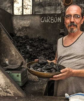 Man in a tank top holding a bowl of charcoal in a dimly lit room with charcoal piles. - Olive Oil Times