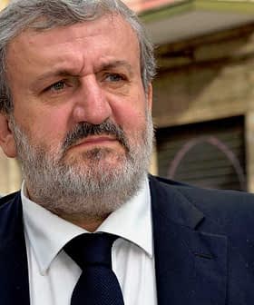 A man with a gray beard wearing a dark suit and tie, looking serious outdoors. - Olive Oil Times