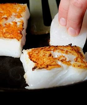 Person placing fish fillets into a skillet while cooking on a stovetop. - Olive Oil Times