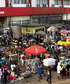 Crowd of people shopping at a bustling outdoor market with various stalls and vehicles. - Olive Oil Times