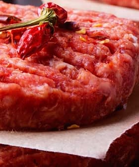 Two raw beef patties stacked on parchment paper with red chili peppers and salt sprinkled around. - Olive Oil Times