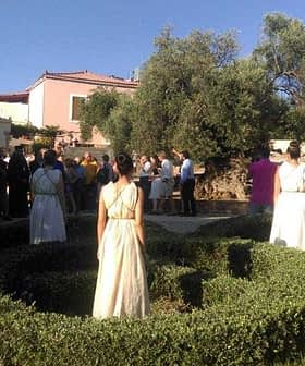A gathering of people outdoors with dancers in white costumes in a garden setting. - Olive Oil Times