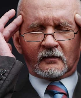 A man with gray hair and glasses holding his head with both hands, appearing deep in thought. - Olive Oil Times