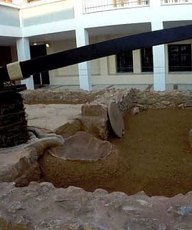 An olive oil pressing setup featuring a large wooden beam and stone base in an outdoor courtyard. - Olive Oil Times