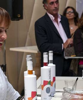 A woman smiling while serving olive oil at a tasting event with attendees in the background. - Olive Oil Times