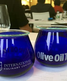 Two blue glass cups labeled 'International Culinary Center' and 'Olive Oil Times' on a table. - Olive Oil Times