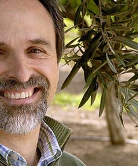 Man with a beard smiling in an olive grove with olive branches in the background. - Olive Oil Times