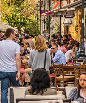 Crowd of people dining at outdoor tables along a city street with pedestrians walking by. - Olive Oil Times