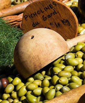 A collection of green olives displayed with wooden utensils and signs indicating prices. - Olive Oil Times