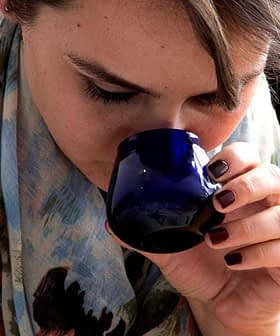 Woman with a scarf smelling from a blue cup while holding a white lid and a pen. - Olive Oil Times