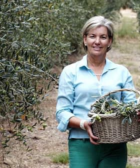 Woman standing in an olive grove, holding a woven basket filled with olives. - Olive Oil Times