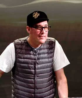 Man wearing a gray vest and black cap standing at a table during a presentation. - Olive Oil Times