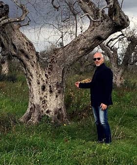 A man in a black jacket standing next to an olive tree in a field. - Olive Oil Times