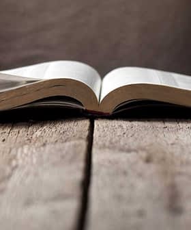 Hands turning pages of an open book resting on a wooden table. - Olive Oil Times