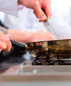 Chef in a white shirt using a frying pan on a gas stove in a kitchen setting. - Olive Oil Times