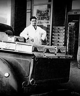 A man in a white coat standing beside a vintage grocery delivery truck filled with boxes of grapes. - Olive Oil Times