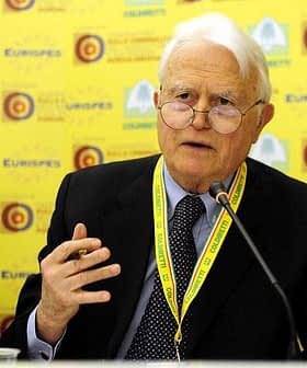 An elderly man with glasses speaking at a conference table with a yellow background. - Olive Oil Times