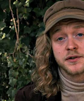 A man with long, curly hair wearing a beige cap and a turtleneck sweater, sitting outdoors. - Olive Oil Times