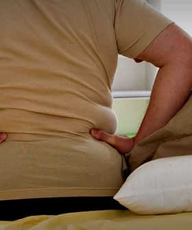 Individual sitting on a bed with hands on lower back, indicating discomfort or pain. - Olive Oil Times
