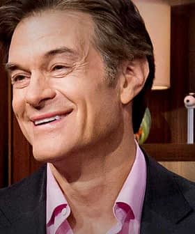 A man with light brown hair smiling while seated in a studio with various objects in the background. - Olive Oil Times