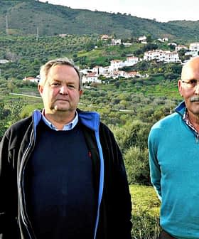 Two men standing side by side in an olive grove with a hillside village in the background. - Olive Oil Times