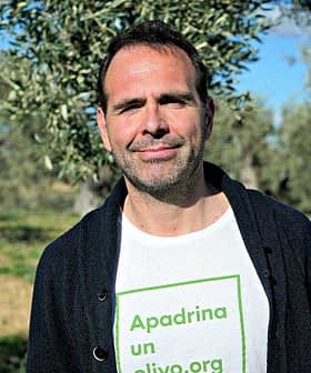 Man standing in an olive grove wearing a white t-shirt with green text. - Olive Oil Times