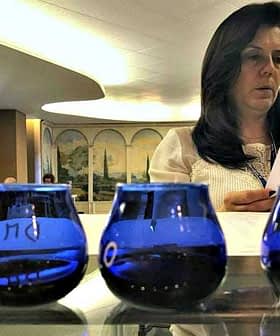 Four blue glass containers arranged on a table with a woman reviewing documents in the background. - Olive Oil Times