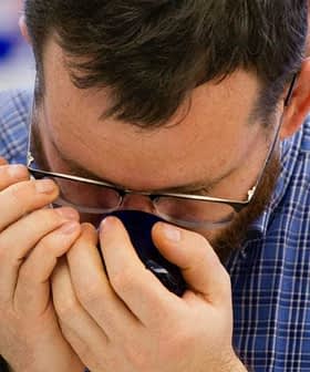 Individual with glasses holding a coffee cup close to their face, inhaling the aroma. - Olive Oil Times