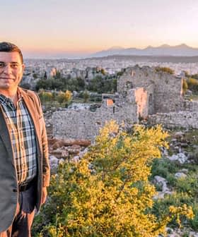 A man wearing a suit stands in front of ancient ruins during sunset with mountains in the distance. - Olive Oil Times