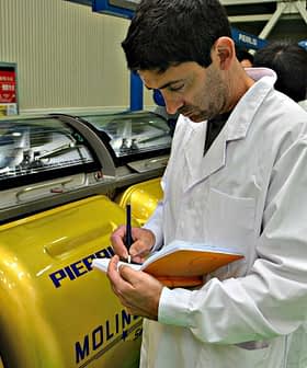 Man in a lab coat taking notes while observing yellow machinery in a facility. - Olive Oil Times