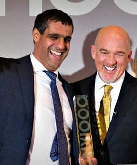Two men smiling and celebrating on stage at an event with a city skyline backdrop. - Olive Oil Times