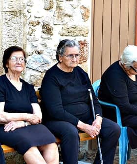 Six elderly women dressed in black and one in pink sitting on a bench outside a building. - Olive Oil Times