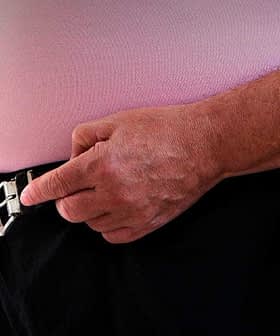 A man adjusting a black belt on his pants with both hands, wearing a pink shirt. - Olive Oil Times
