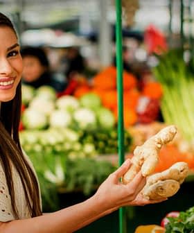 A woman smiling while holding a piece of ginger in a market filled with fresh produce. - Olive Oil Times