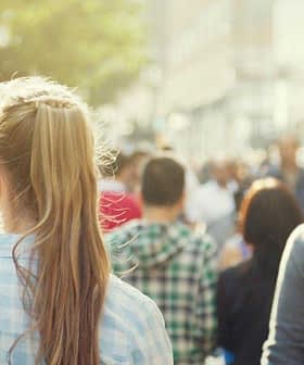 A view of a busy street with people walking in various directions, including a woman with long hair in a plaid shirt. - Olive Oil Times