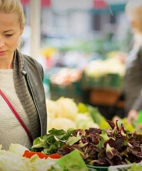 Woman examining fresh vegetables at a market stall surrounded by other shoppers. - Olive Oil Times
