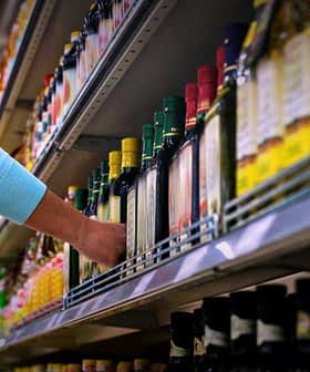 Individual reaching for a bottle of olive oil on a grocery store shelf with various products. - Olive Oil Times