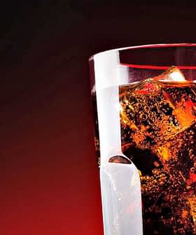 A glass filled with cola and ice cubes against a dark background. - Olive Oil Times