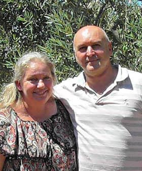 A couple standing together in an olive grove, smiling at the camera. - Olive Oil Times