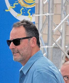 Man with short dark hair wearing sunglasses and a blue shirt, standing in front of a blue background. - Olive Oil Times