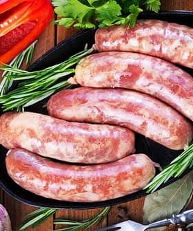 Five raw sausages arranged on a black platter with herbs and garlic nearby. - Olive Oil Times