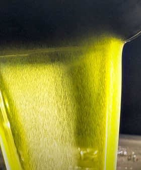 Stream of green olive oil being poured from a metal container into a stainless steel surface. - Olive Oil Times