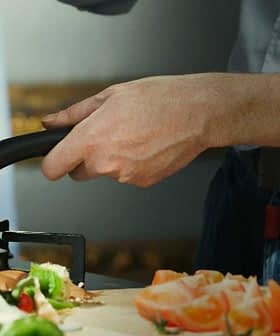 A person stirring food in a frying pan while preparing ingredients on a kitchen countertop. - Olive Oil Times