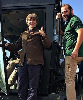 Two men standing on an olive harvesting machine, one giving a thumbs up while the other stands beside him. - Olive Oil Times