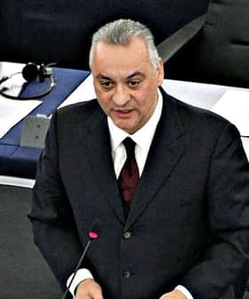 A man speaking at a podium in a legislative assembly with numbered seats visible in the background. - Olive Oil Times