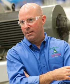 Man wearing a blue shirt and safety glasses, gesturing while speaking in an industrial setting. - Olive Oil Times