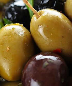 A bowl containing a variety of green and black olives garnished with herbs. - Olive Oil Times