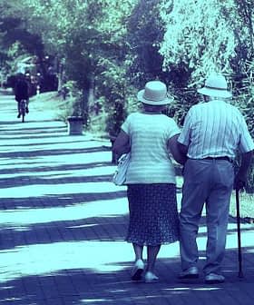 An elderly couple walking together on a tree-lined path, both wearing hats and holding hands. - Olive Oil Times