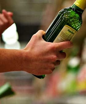 A person holding a green bottle of olive oil while shopping in a grocery store aisle. - Olive Oil Times
