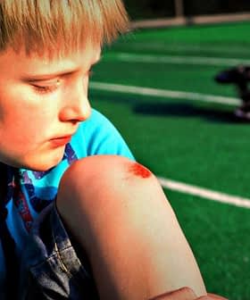 Young boy sitting on a green field with a scraped knee, looking down at the injury. - Olive Oil Times
